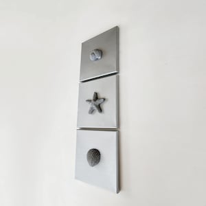 Op de afbeelding: Drie vierkante, zilverkleurige wandkunstwerken. Elk stuk heeft een getextureerde, grijze zeeschelp, zeester of spiraalschelp. De kunst is op een witte muur gemonteerd en creëert een kustthema.