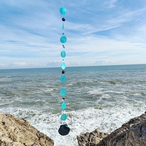 Puede incluir: Un móvil turquesa y negro con piezas redondas y con forma de concha colgando de una cuerda. El móvil está colgando frente a un océano azul y una costa rocosa.