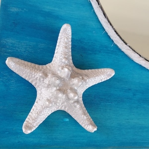A white starfish with a textured surface is attached to a turquoise blue wood background. It is a part of a nautical style decorative mirror.
