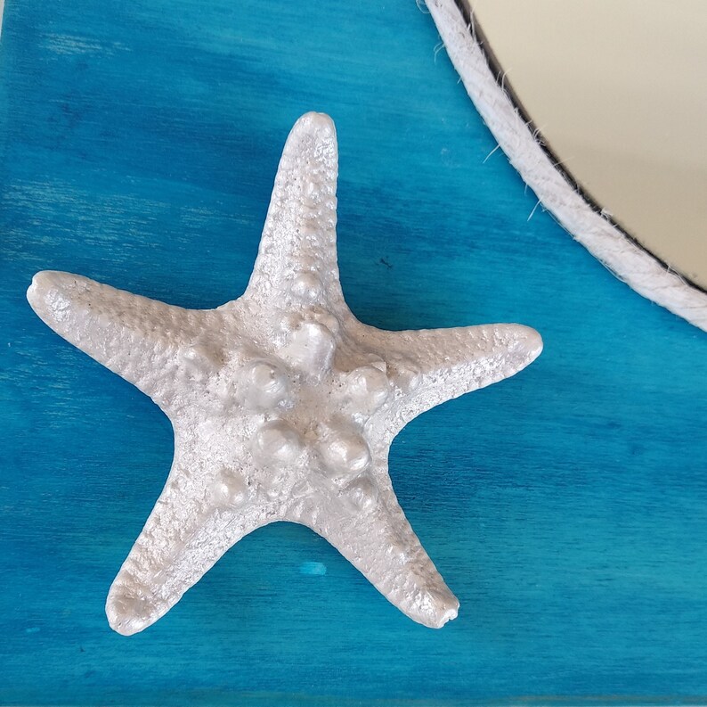A white starfish with a textured surface is attached to a turquoise blue wood background. It is a part of a nautical style decorative mirror.