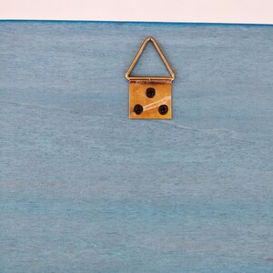 Closeup of a light blue wooden surface with a gold colored triangular hanging hook attached to it. The hook has three small black screws.