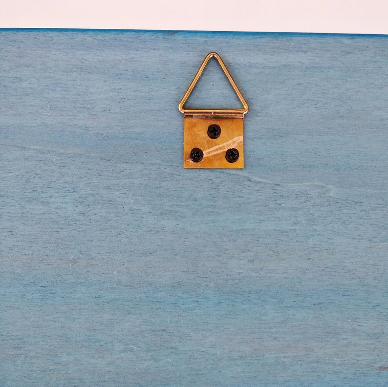 Closeup of a light blue wooden surface with a gold colored triangular hanging hook attached to it. The hook has three small black screws.