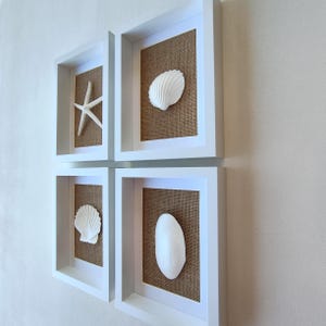 Peut inclure: Un ensemble de quatre cadres carrés blancs de type boîte vitrine disposés en grille. Chaque cadre contient un fond en toile de jute marron naturel et un coquillage ou une étoile de mer blanc, incluant une étoile et des coquillages.