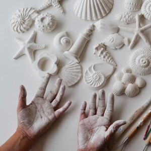 Una colección de adornos blancos de conchas y estrellas de mar esculpidas. Se exhiben figuras de diversas formas y tamaños, incluyendo una estrella de mar, una concha espiral y otras formas marinas. Se aprecian las manos del artista, cubiertas con material blanco, junto con sus herramientas de escultura.