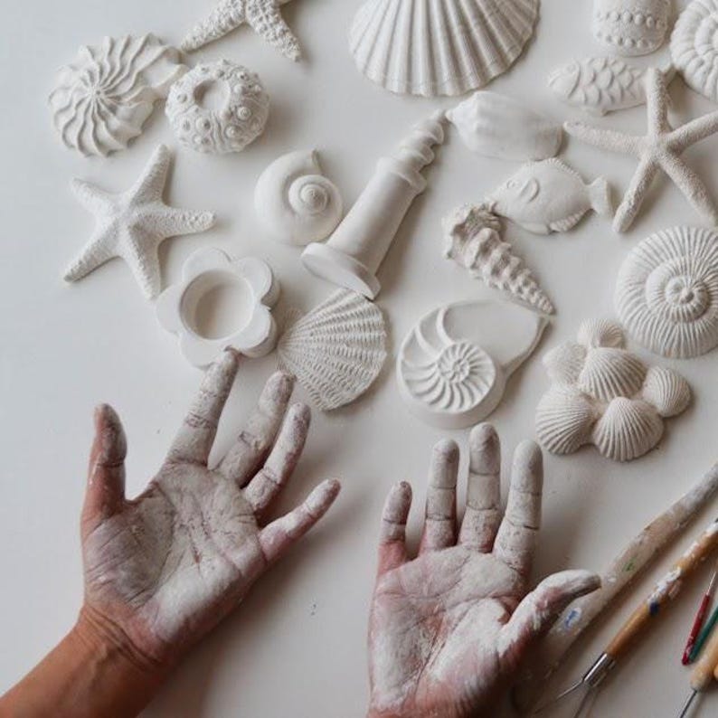 Una colección de adornos blancos de conchas y estrellas de mar esculpidas. Se exhiben figuras de diversas formas y tamaños, incluyendo una estrella de mar, una concha espiral y otras formas marinas. Se aprecian las manos del artista, cubiertas con material blanco, junto con sus herramientas de escultura.
