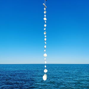 Può includere: Un mobile decorativo di conchiglie bianche è appeso contro un cielo azzurro e il mare. Le conchiglie, di varie dimensioni, sono infilate insieme. L'orizzonte dell'oceano è visibile sullo sfondo, creando una serena scena costiera.