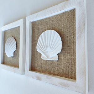 Op de afbeelding: Twee ingelijste wandkunstwerken met witte schelpen. De schelpen zijn gemonteerd op een jute-achtige achtergrond in witte houten frames. De frames hebben een verweerde, rustieke uitstraling, wat het kustthema versterkt.