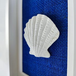 Una escultura de concha marina blanca en relieve y texturizada que posa sobre una tela de arpillera en azul dentro de un marco blanco, obra de arte de playa.