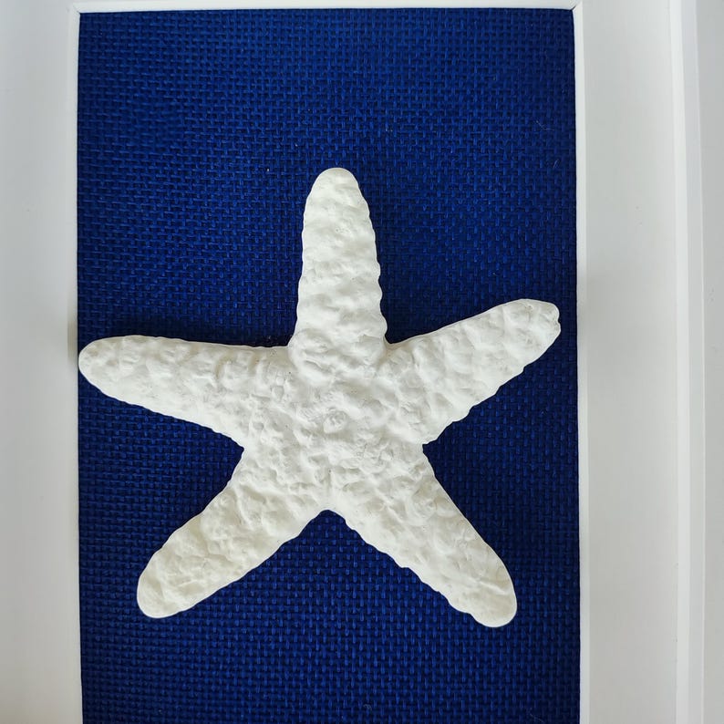 Una estrella de mar de cinco dedos de cerámica blanca montada sobre un fondo azul en un marco blanco. Objeto de decoración costera.