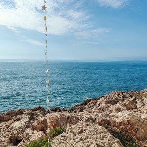 May include: A white string of decorative seashells and stars hangs from the top of a rocky cliff overlooking a blue ocean. The sky is blue with white clouds.
