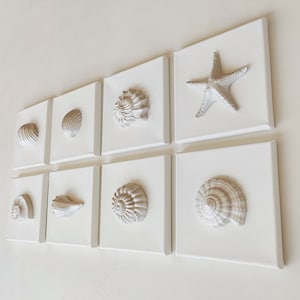 Peut inclure: Un ensemble de neuf tableaux muraux en toile blanche représentant différents coquillages et une étoile de mer. Les coquillages sont de formes et de tailles variées, et l'étoile de mer est au centre de la rangée du haut.