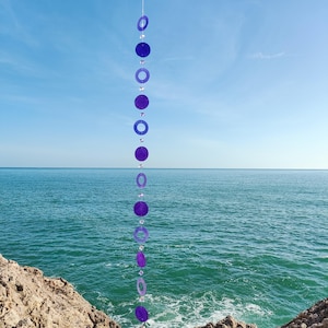 Puede incluir: Móvil colgante vertical con círculos y anillos morados alternados, intercalados con cuentas transparentes. El móvil está suspendido sobre un fondo de cielo azul y mar. Los círculos y anillos son de diferentes tamaños.