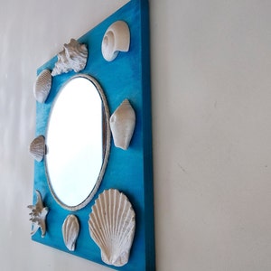 A decorative turquoise blue wooden framed mirror with a round mirror surrounded by white shells. The frame is painted bright blue and the shells are arranged in a circle around the mirror. Coastal wall decor.