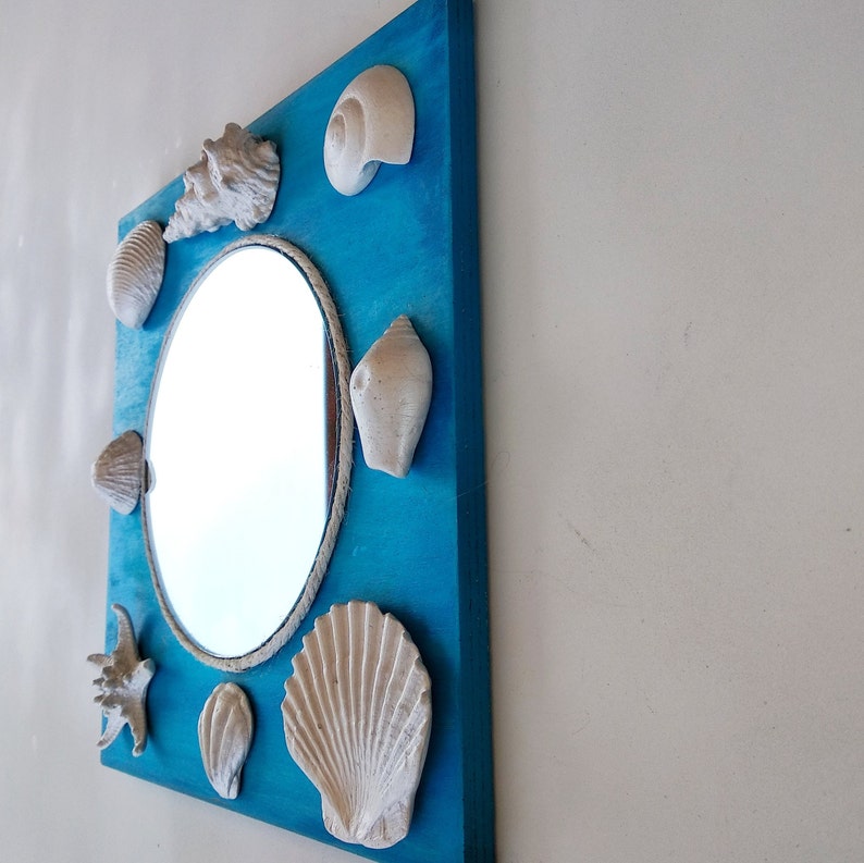 A decorative turquoise blue wooden framed mirror with a round mirror surrounded by white shells. The frame is painted bright blue and the shells are arranged in a circle around the mirror. Coastal wall decor.