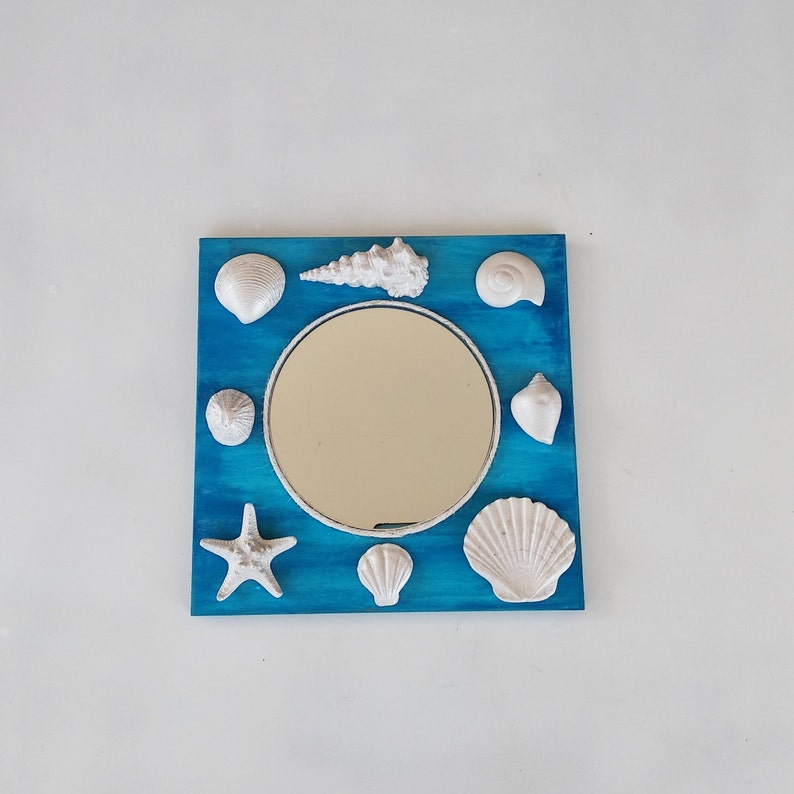 A blue square wooden mirror decorated with white seashells. The round mirror glass is surrounded by white seashell sculptures of various shapes and sizes.