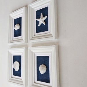 May include: Four white picture frames with blue mats and white seashells and a starfish. The frames are arranged in a grid pattern on a white wall.