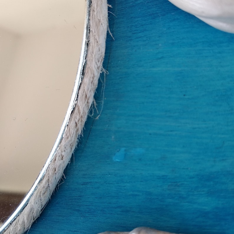Closeup of a round mirror with a white rope trim. The mirror is placed on a bright turquoise blue background. It is a coastal style decorative mirror.