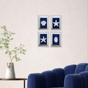 Cuatro obras de arte enmarcadas en blanco con fondo azul, cada una con una concha o estrella de mar blanca diferente. Las imágenes están dispuestas en un cuadrado en una pared blanca.
