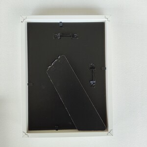 Parte trasera de un marco de fotos blanco con un caballete negro. El marco tiene un gancho para poderlo colgar en la pared.