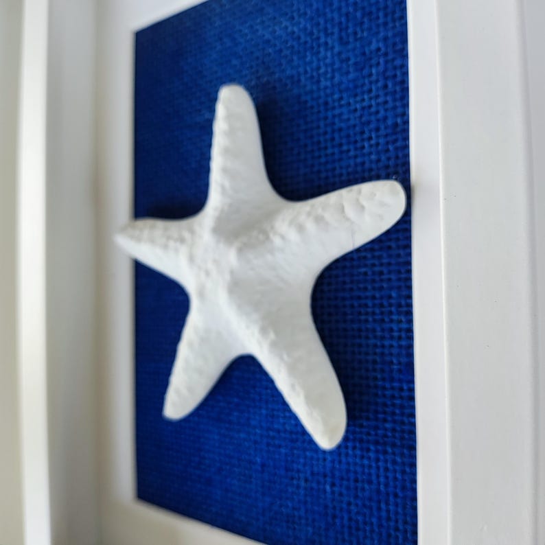 Una escultura de una estrella de mar blanca que posa sobre un tejido de yute azul marino, dentro de un marco blanco. Obra de arte de playa.