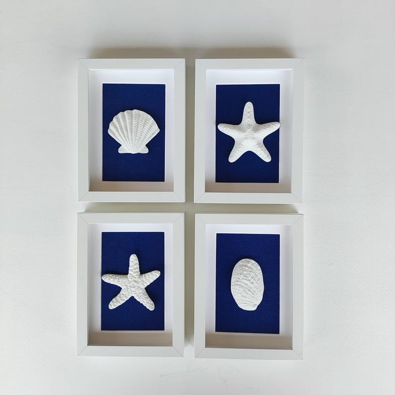 Cuatro cuadros decorativos enmarcados con imágenes blancas con fondos azules. Cada imagen presenta una concha o una estrella de mar blanca en relieve. Decoración con estilo costero.