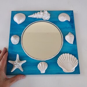 A blue square wooden frame with a round mirror in the center. The frame is decorated with white shells of various shapes and sizes that surround the mirror. Nautical decor.