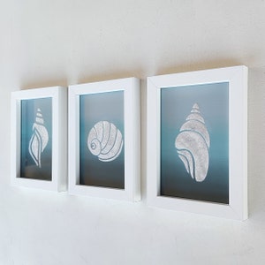 Könnte beinhalten: Drei weiße Bilderrahmen mit blauem Hintergrund und weißen Muschelmotiven. Die Muscheln sind in verschiedenen Stilen.