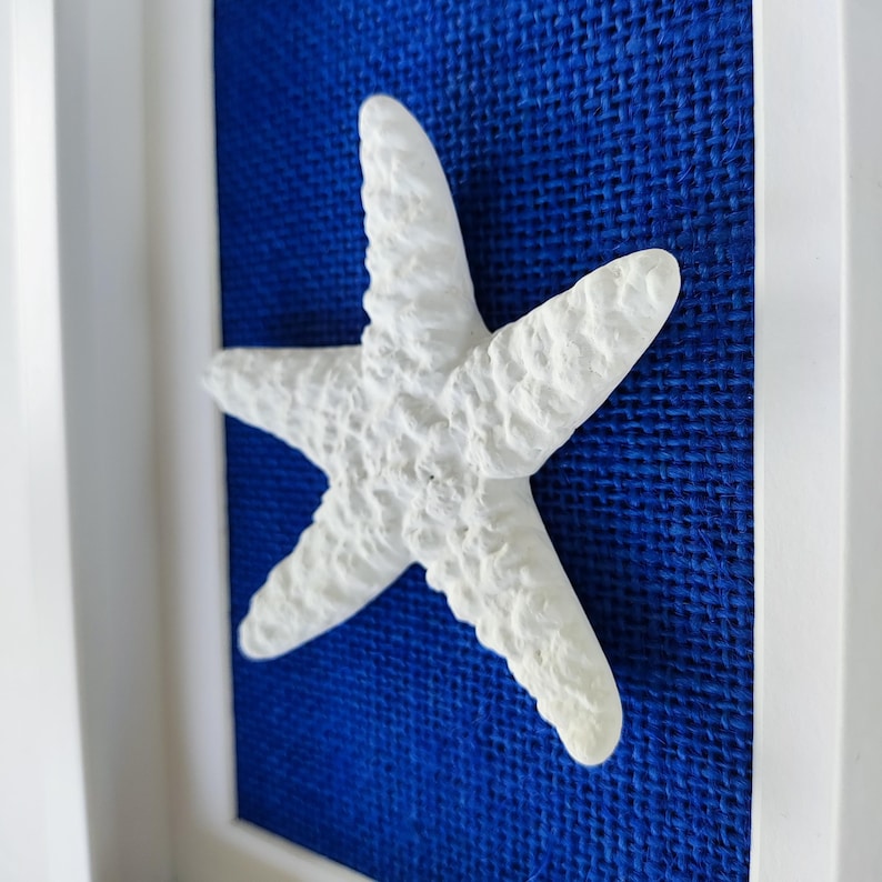 Una pequeña muy realista escultura de una estrella de mar blanca que posa sobre un tejido de yute azul marino, dentro de un marco blanco. Obra de arte costero.