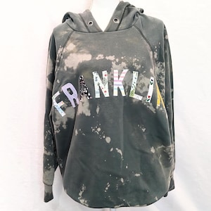 Peut inclure: Un sweat-shirt à capuche vert avec un motif éclaboussé de javel. Le mot "FRANKLIN" est écrit en patchs de tissu colorés.