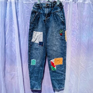 May include: Blue denim jeans with colorful fabric patches sewn on the front. The patches include a plaid pattern, a green and white checkered pattern, and a red and yellow pattern.