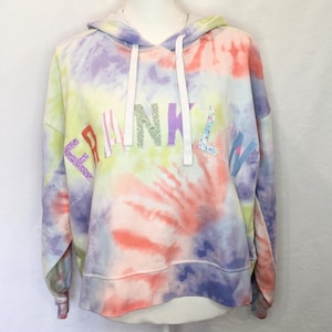 Op de afbeelding: Een tie-dye hoodie in een werveling van kleuren, waaronder geel, paars, roze en blauw. Het woord "FEMININE" staat in verschillende decoratieve lettertypen over de borst. De hoodie heeft een capuchon met trekkoord en lange mouwen.