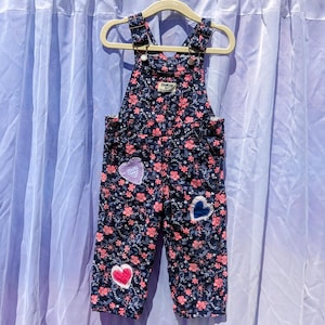 May include: Blue denim dungarees with a pink floral print and three heart-shaped patches. The dungarees have adjustable straps and a button closure.