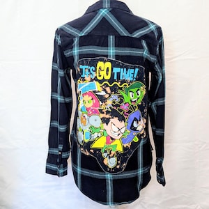 May include: A long-sleeved, button-down shirt with a black and teal plaid pattern. The back features a colorful graphic of cartoon characters with the text "IT'S GO TIME!"