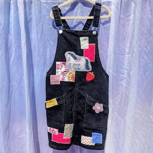 May include: Black denim overall dress with colorful patchwork details, including a pink flower, a strawberry, and a white horse.