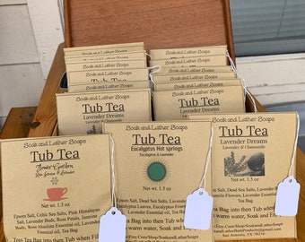 Può includere: Una scatola di legno piena di piccoli sacchetti di carta marrone contenenti miscele di tè da bagno. I sacchetti sono etichettati con il nome "Soak and Lather Soaps" e il tipo di tè, come "Tub Tea Flower Garden", "Tub Tea Lavender Dreams" e "Tub Tea Eucalyptus Hot Springs". Ogni sacchetto contiene 1,5 once di tè da bagno e include un elenco di ingredienti e istruzioni per l'uso.