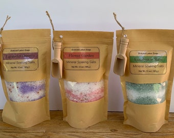 May include: Three brown paper bags with scoops attached, each containing mineral soaking salts. The bags are labeled with the scents "Lavender Dreams", "Flower Garden", and "Eucalyptus Rain". Each bag contains 12 ounces (340 grams) of salts.