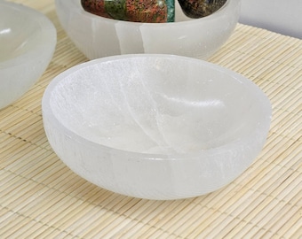 May include: Two white selenite bowls, one with a variety of green, red, and brown stones inside. The bowls are on a light brown bamboo mat.