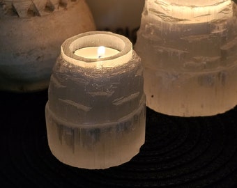 Può includere: Due portacandele cilindrici realizzati in cristallo di selenite bianco traslucido con una consistenza esterna ruvida e naturale. Il portacandele più piccolo contiene un lumino acceso, mentre quello più grande è posizionato dietro.