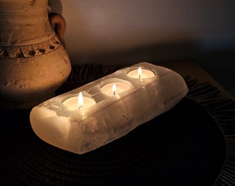 Può includere: Un portacandele rettangolare in selenite bianca con tre lumini accesi. Il portacandele è appoggiato su una superficie scura, con un vaso beige strutturato sullo sfondo. Le candele emettono un bagliore caldo.