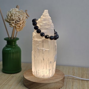 Könnte beinhalten: Eine weiße Selenit-Kristalllampe mit Holzsockel, von innen beleuchtet. Ein dunkelblaues Perlenarmband ist über dem Kristall drapiert. Links befindet sich eine grüne Vase mit einer dekorativen Blume. Die Lampe ist etwa 20 cm hoch.
