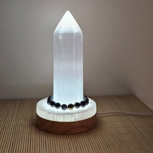Op de afbeelding: Een witte selenietkristal lamp met een houten voet en een zwarte kralen armband erop. De lamp is aan en geeft een warm licht.
