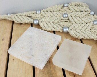 May include: Two square, pale stone coasters. One is a larger, textured square, and the other is a smaller, smooth square. The coasters are displayed on a wooden surface, with a macrame decoration and silver beads in the background.
