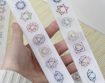 Könnte beinhalten: Weiße Selenit-Stäbe mit bunten Chakra-Symbolen. Jeder Stab zeigt sieben Symbole, die jeweils ein Chakra repräsentieren. Die Stäbe werden in einer Hand gehalten, wodurch die detaillierten Designs und die natürliche Textur des Selenits hervorgehoben werden.