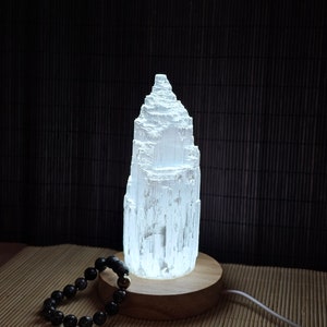 Könnte beinhalten: Eine weiße Selenit-Kristall-Turmlampe auf einem Holzsockel mit einem schwarzen Perlenarmband im Vordergrund. Die Lampe ist beleuchtet und strahlt ein warmes Licht aus.