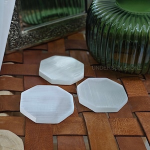 Può includere: Tre sottobicchieri in selenite bianca a forma esagonale, appoggiati su una superficie in pelle marrone. I sottobicchieri sono lisci e hanno una lucentezza delicata.