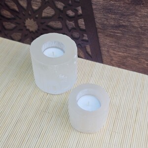 Pu&ograve; includere: Due portacandele in selenite bianca, uno pi&ugrave; grande dell'altro, con una piccola candela da t&egrave; bianca all'interno di ciascuno. I portacandele sono su un tappetino di bamb&ugrave; marrone chiaro.