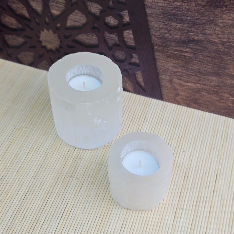 Pu&ograve; includere: Due portacandele in selenite bianca, uno pi&ugrave; grande dell'altro, con una piccola candela da t&egrave; bianca all'interno di ciascuno. I portacandele sono su un tappetino di bamb&ugrave; marrone chiaro.