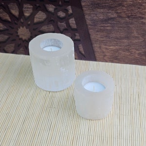 Pu&ograve; includere: Due portacandele in selenite bianca con forma rotonda e cilindrica. I portacandele sono su un tappetino di bamb&ugrave; marrone chiaro.
