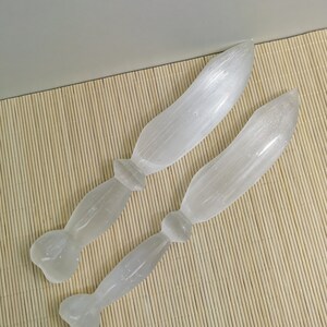 Könnte beinhalten: Zwei weiße Selenit-Massage-Tools werden auf einer hellbraunen Bambusmatte gezeigt. Die Tools haben eine glatte, längliche Form mit einem abgerundeten Ende und einem Griff. Der Selenit hat ein durchscheinendes Aussehen.