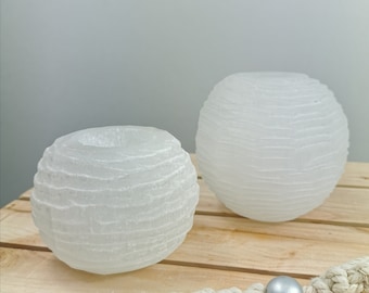 Può includere: Due portacandele sferici bianchi con un design strutturato a strati. Uno ha un'apertura centrale per una candela, l'altro è una sfera piena. Sono su una superficie di legno con elementi decorativi.